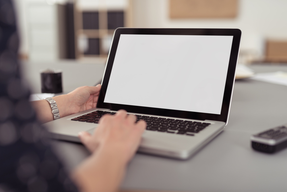 Businesswoman sitting at her desk navigating the internet on a laptop computer using the trackpad, over the shoulder view of the blank screen Businesswoman sitting at her desk navigating the internet on a laptop computer using the trackpad, over the shoulder view of the blank screen